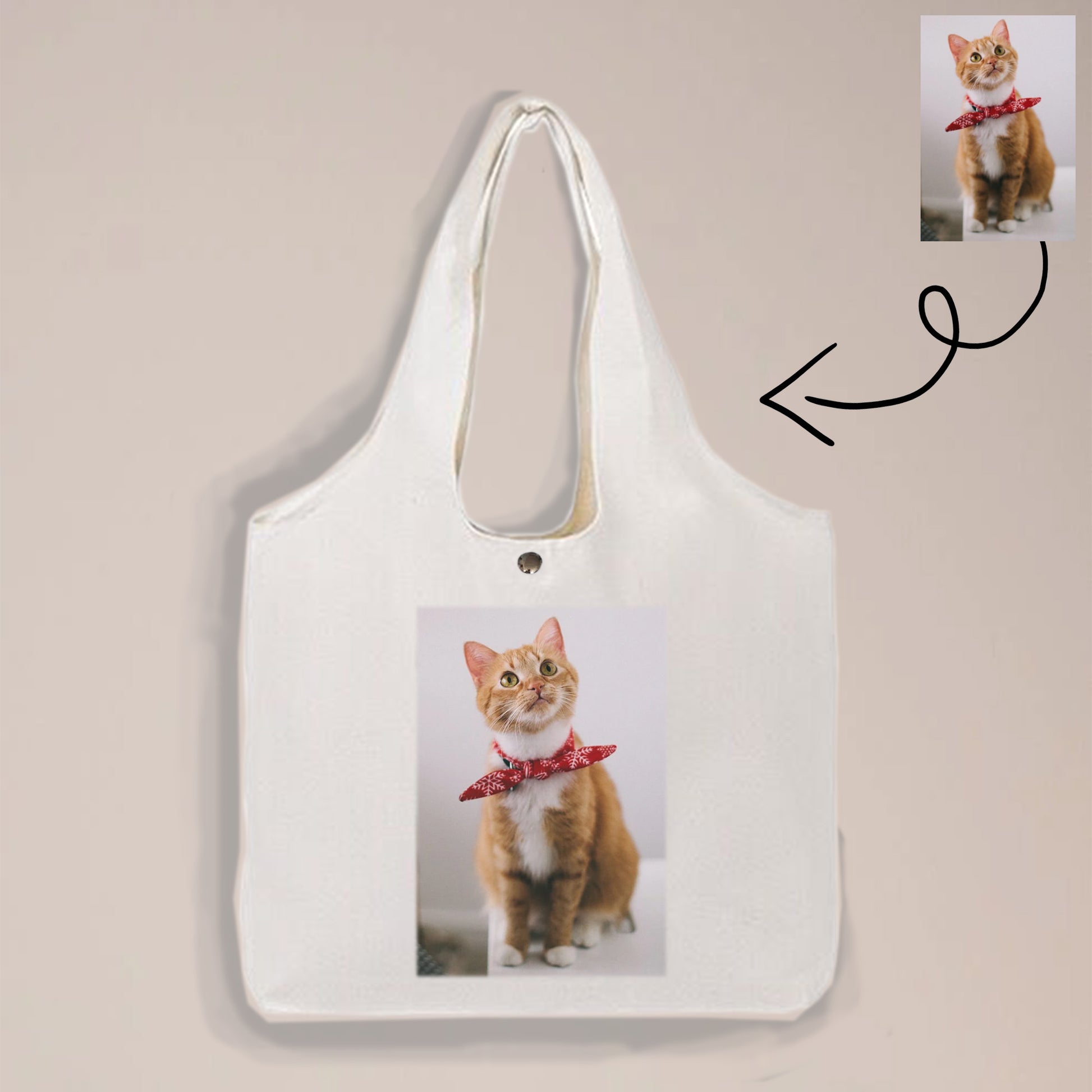 Image of a white tote bag with a picture of a cat on it. The cat is wearing a red bow and sitting on the floor.