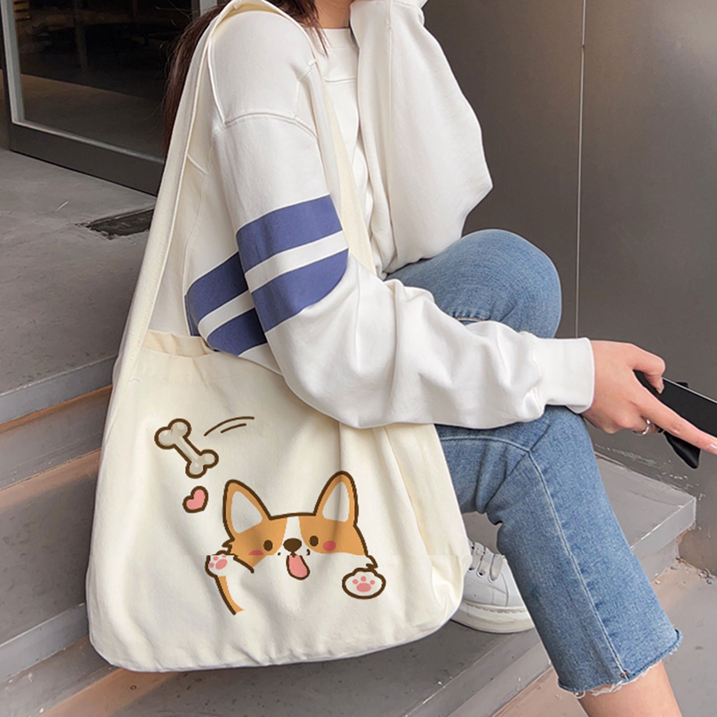 A person sitting on the stairs in blue jeans and white sneakers is wearing a personalized white crossbody bag with a cute cat, bone, and red heart printed on it.